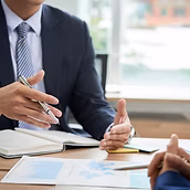 Symbolbild Beratung. Zwei Menschen sitzen sich an einem Tisch gegenüber. Zwischen ihnen liegen Unterlagen. Eine Person spricht und gestikuliert, von der anderen Person sind nur die gefalteten Hände zu sehen.